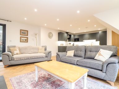 An open-plan living room with two grey sofas a wooden coffee table a patterned rug kitchen cabinets and appliances at Wildwood in Newquay