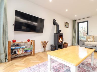 A living room with a wall mounted television wood burning stove wooden coffee table and a grey sofa at Wildwood in Newquay