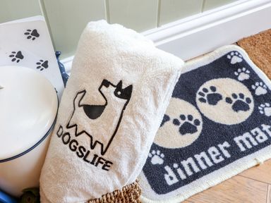 A white towel with a dog graphic and the word dogslife next to a feeding mat with paw prints and the words dinner mat on a wooden floor at Wildwood in Newquay