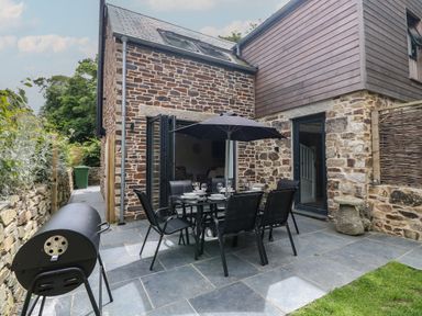 An outdoor patio with a dining table and chairs under an umbrella next to a stone house with glass doors and a barbecue grill at Wildwood in Newquay