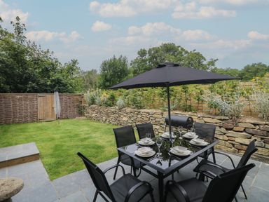An outdoor patio with a black dining table and six chairs under a black umbrella set with plates and glasses next to a stone wall and green lawn at Wildwood in Newquay