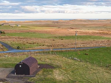 A small curved black building on a grassy field near a road with fields and ocean in the background at Heisgeir in Sollas Isle Of North Uist
