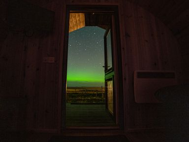 A wooden cabin interior with a door open to a starry night sky and green northern lights at Heisgeir in Sollas Isle Of North Uist