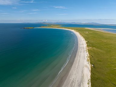 A curved sandy beach next to green grassy land with distant islands over blue sea at Heisgeir Sollas Isle Of North Uist