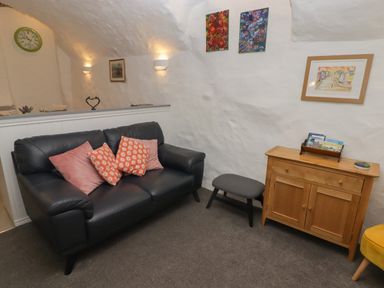 A living room with a black leather sofa and colorful cushions wooden cabinet and framed pictures on white walls at Le Durkestrete Cellar in Haverfordwest