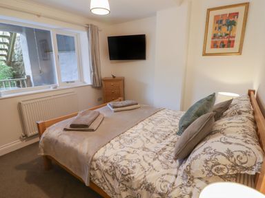 A bedroom with a bed featuring patterned bedding and pillows a wall-mounted TV a window with curtains and a small wooden cabinet at Le Durkestrete Cellar in Haverfordwest