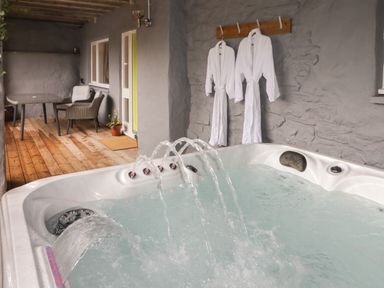 An outdoor covered patio with a hot tub with water jets a wooden deck table and chairs two white robes hanging on a wall at Le Durkestrete Cellar in Haverfordwest