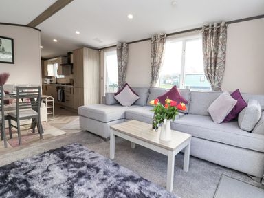 A living room with a gray sectional sofa and a coffee table with flowers near a kitchen and dining area at Ginger Fox Lodge in East Heslerton near Cayton