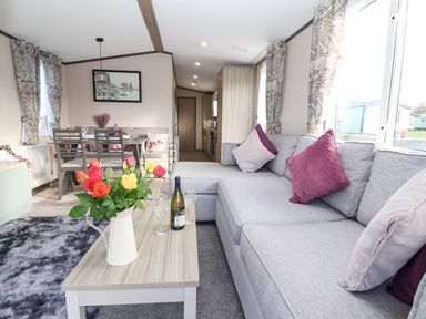 A living room with a gray sofa, a wooden coffee table with flowers and wine, and a dining table with chairs at Ginger Fox Lodge in East Heslerton near Cayton