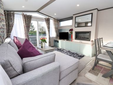 A living room with a gray sectional sofa with purple cushions a coffee table with flowers a TV on a cabinet and dining table with chairs at Ginger Fox Lodge in East Heslerton near Cayton