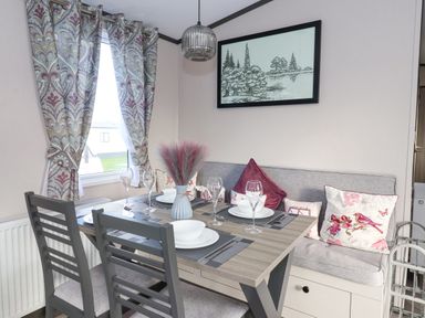 A dining area with a table set with bowls plates and wine glasses two gray chairs a cushioned bench with pillows curtains on the window and a framed drawing of trees on the wall at Ginger Fox Lodge in East Heslerton near Cayton
