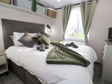 A bedroom with a bed with pillows and folded towels near a window with curtains and a dresser with a TV at Ginger Fox Lodge in East Heslerton near Cayton