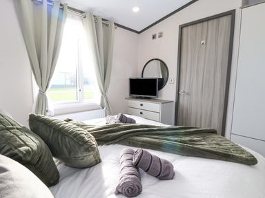 A bedroom with a bed with green pillows and towels in front of a window with curtains a dresser with a television and a round mirror and a closed wooden door at Ginger Fox Lodge in East Heslerton near Cayton