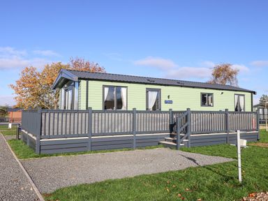 A single story mobile home with a gray deck and a gravel driveway surrounded by grass at Ginger Fox Lodge in East Heslerton near Cayton