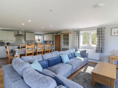 An open living room and kitchen with a large gray sectional sofa, wooden dining table with chairs, and kitchen cabinets at Curtau in Llanfaredd near Builth Wells