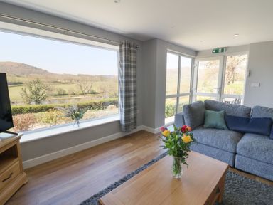 A living room with a grey sofa a wooden coffee table with flowers and large windows showing a countryside view at Curtau in Llanfaredd near Builth Wells