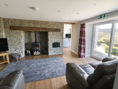 A living room with stone fireplace and wood-burning stove a gray rug leather sofas and large sliding glass doors at Curtau Llanfaredd near Builth Wells