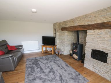 A living room with a gray sofa a wooden TV stand a flat screen TV a stone fireplace with a wood burner and a gray rug on a wooden floor at Curtau in Llanfaredd near Builth Wells