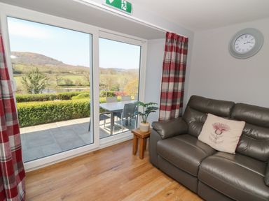A living room with a leather sofa a side table with a plant curtains and a large window showing an outdoor dining area and a countryside view at Curtau in Llanfaredd near Builth Wells