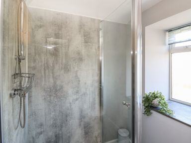 A shower area with a glass door a showerhead on a grey textured wall and a window with a small plant on the sill at Curtau in Llanfaredd near Builth Wells