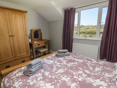A bedroom with a bed covered with floral patterned bedding wooden wardrobe dressing table and window with purple curtains showing a countryside view at Curtau in Llanfaredd near Builth Wells