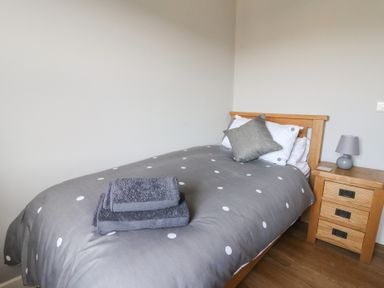 A single bed with gray polka dot bedding and folded towels next to a wooden bedside table with a lamp at Curtau Llanfaredd near Builth Wells