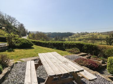 A wooden picnic table on a gravel area with a lawn and hedges overlooking fields and trees at Curtau in Llanfaredd near Builth Wells