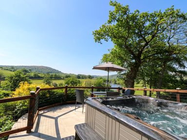 An outdoor patio with a hot tub table and chairs with an umbrella overlooking a river and green hills at Curtau in Llanfaredd near Builth Wells
