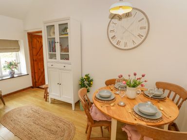 A dining area with a wooden round table set for four with pink flowers in a vase and a white cabinet with glass doors at The Stables Near Solva