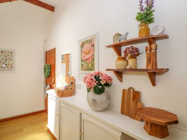 A white sideboard with a vase of pink flowers wooden cutting boards and a wooden shelf with jars and a small plant at The Stables Near Solva