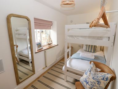 A bedroom with white bunk beds a wooden chair with a cushion a window with a model boat and a framed picture a full length mirror and striped rug at The Stables Near Solva
