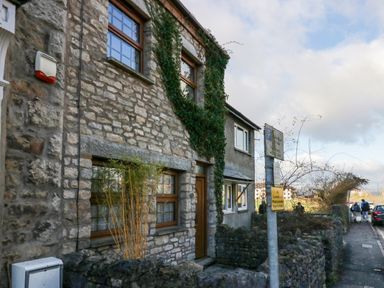 Exterior of a stone house with wooden windows and a wooden door on a street with parked cars and a parking sign in Arnside
