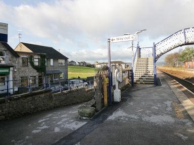 A railway platform with a footbridge and way out sign near houses and a grassy field at Time and Tide in Arnside