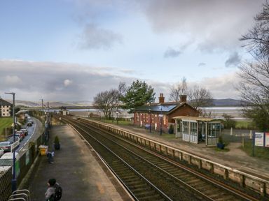 A railway station with two tracks and platforms a small brick building and a glass shelter with a road and parked cars nearby at Time and Tide in Arnside