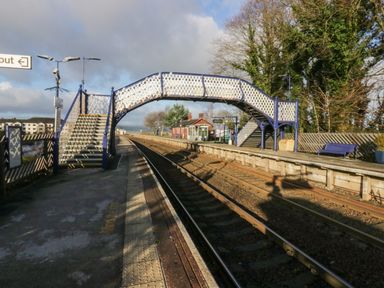 A railway station platform with a blue and white footbridge over the tracks surrounded by trees and buildings at Time and Tide in Arnside
