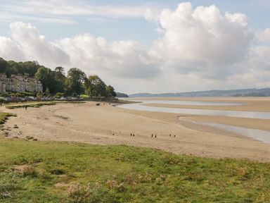 A sandy beach with tidal pools and grass in the foreground and houses and trees along the shore at Time and Tide in Arnside