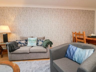 A living room with a gray sofa with cushions and throw blankets a gray armchair with a blue cushion a wooden dining table with chairs and a lamp on a side table at Time and Tide in Arnside