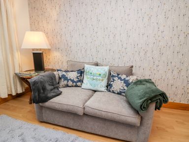 A small sofa with cushions and blankets next to a side table with a lamp in a room with floral wallpaper at Time and Tide in Arnside