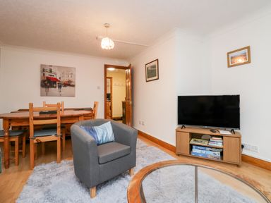 A living room with a gray armchair a wooden dining table a TV on a wooden stand and paintings on the wall at Time and Tide in Arnside