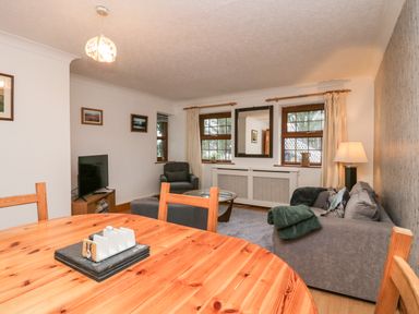 A living room with a wooden dining table chairs a sofa armchair a coffee table a TV and three windows at Time and Tide in Arnside