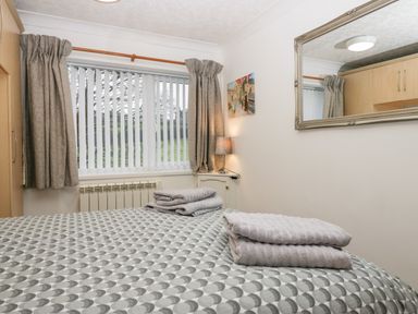 A bedroom with a bed covered in patterned bedding and towels folded on top a window with vertical blinds and curtains a lamp on a small table and a large mirror on the wall at Time and Tide in Arnside