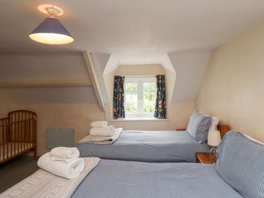 A bedroom with two single beds with blue checkered bedding a crib and a window with floral curtains at Cherry Tree Cottage in Fallodon near Embleton