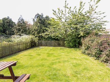 A backyard with a wooden picnic table green grass a wooden fence and trees at Cherry Tree Cottage in Fallodon near Embleton