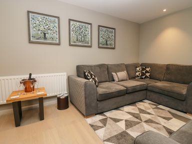 A living room with a grey sectional sofa a patterned rug three framed pictures on the wall and a small wooden table with wine glasses and a wine cooler at Brimstone in Backbarrow near Ulverston