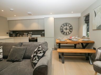 An open plan kitchen and dining area with a grey sofa in the foreground a wooden dining table with benches and wall clock on the wall at Brimstone in Backbarrow near Ulverston