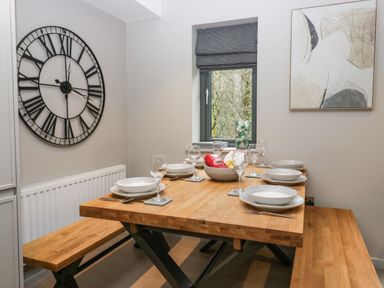 A dining area with wooden table and benches set with plates glasses and cutlery a large clock on the wall and a window with a patterned blind at Brimstone in Backbarrow near Ulverston