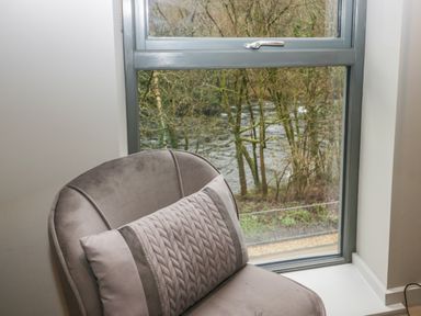 A cushioned chair with a pillow in front of a window showing trees and a river outside at Brimstone Backbarrow near Ulverston