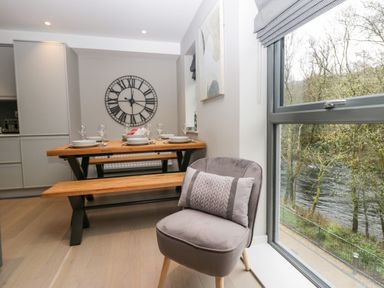 A dining area with a wooden table set with dishes and glasses a wooden bench a gray cushioned chair with a pillow and a large window showing trees by a river at Brimstone in Backbarrow near Ulverston