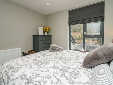 A bedroom with a bed covered in patterned bedding a gray pillow folded towels on the bed a dresser with a vase of sunflowers and a window with gray blinds at Brimstone in Backbarrow near Ulverston