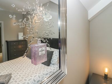 A bedroom with a bed a patterned duvet and pillows a chest of drawers and a framed mirror with a perfume bottle design at Brimstone in Backbarrow near Ulverston